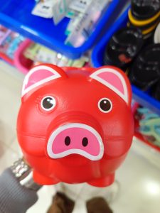 A bright red piggy bank with large, cartoonish eyes and pink accents on its ears and snout is held up against a blurred background of colorful storage bins and items. 