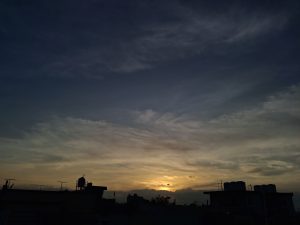 A warm golden sunset spreads across the horizon, casting soft light through layers of wispy clouds, while the dark silhouettes of rooftops, water tanks, and antennas stand in quiet contrast against the vast, darkening sky.