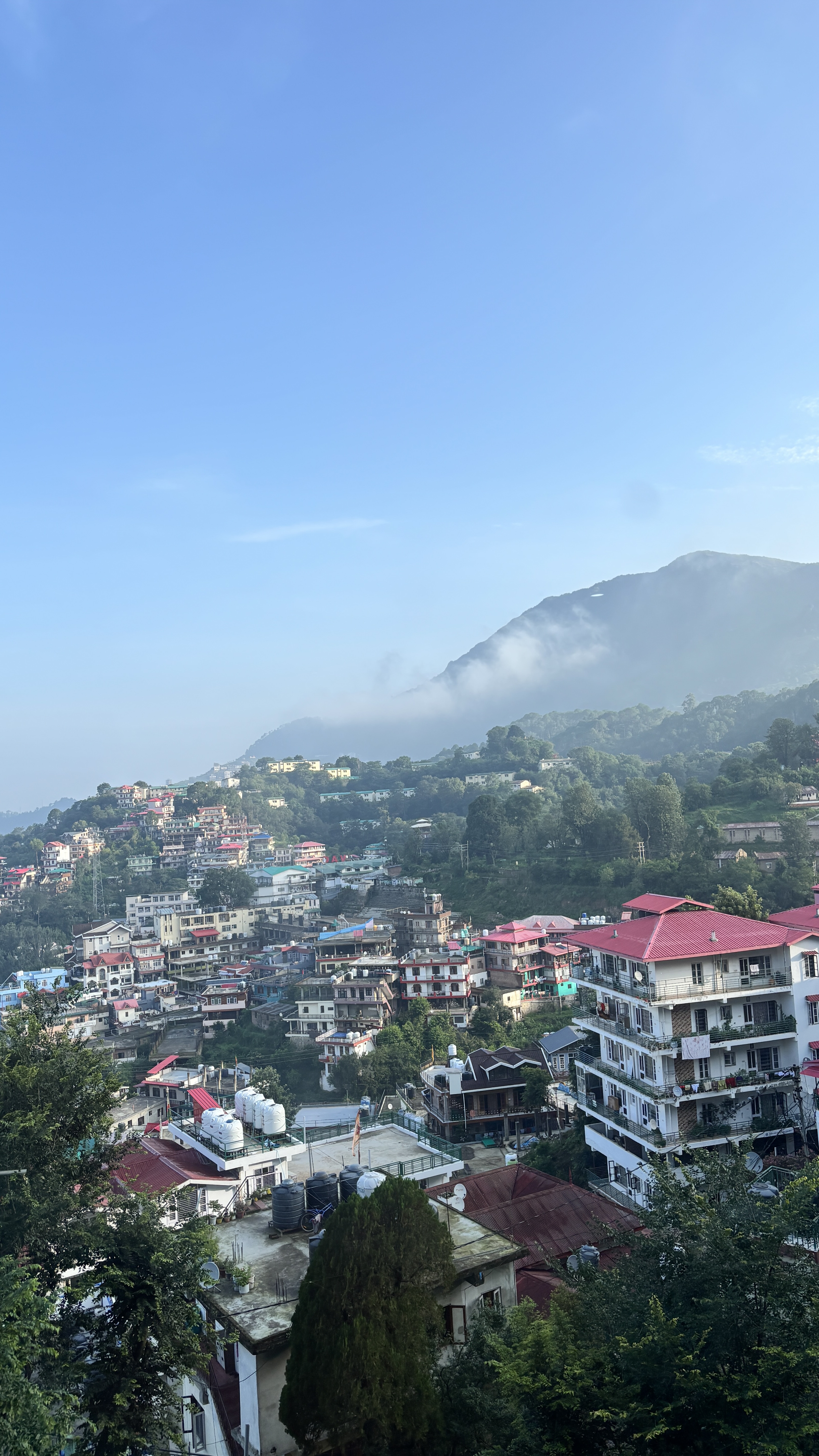 The picture presents mist or low clouds are drifting across the side of the mountain, giving it a soft, dreamy appearance. The buildings are tightly packed and built along steep slopes, creating layred look. Most of the houses have bright coloured roof and the town is surrounded by dense trees and forested hills.
