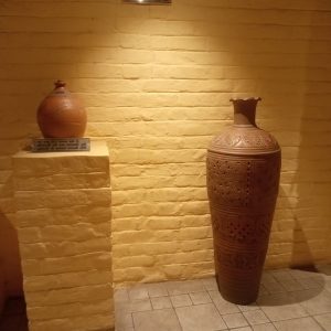 Two beautifully carved terracotta pots against a yellow brick wall, showcasing traditional Indian pottery—one proudly displayed with a plaque noting its 50+ years of cultural heritage