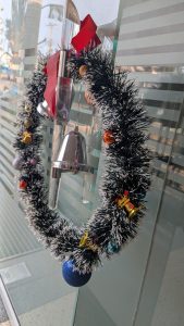 A decorative Christmas wreath made of black and silver tinsel, adorned with colorful ornaments, including small bells, glittery balls, and ribbons.