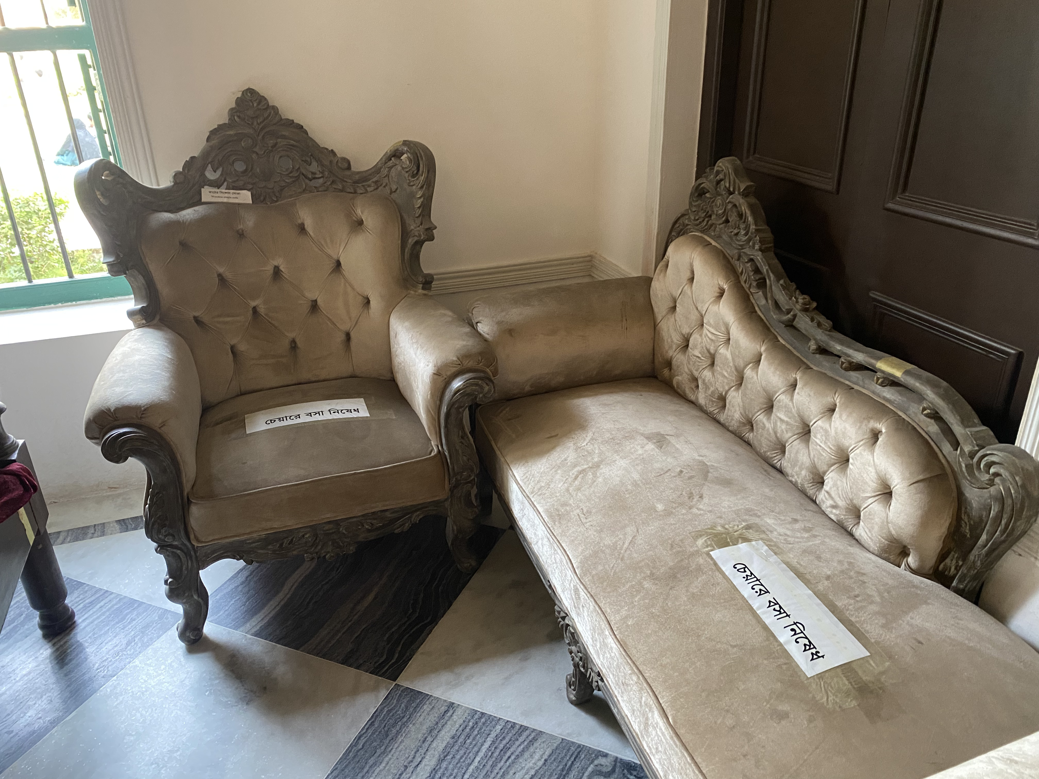 Ornate beige armchair and sofa with tufted upholstery, placed in the corner of a room with tiled floor. Labels in bengali prohibit sitting on the furniture.
