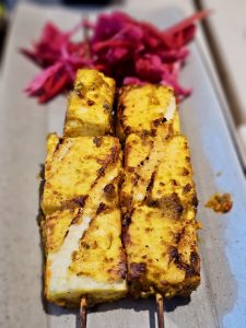 Golden grilled paneer tikka pieces served on skewers with spices and light char marks. The close framing highlights texture and color, captured at a restaurant in Kozhikode, Kerala, showcasing rich vegetarian cuisine. 
