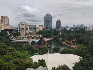 The image shows a cityscape with tall modern buildings overlooking a large water-themed amusement park surrounded by lush greenery. There are pools, slides and decorative rock structures, with a calm lake in the center and dense trees in the foreground in Bandar Sunway, Malaysia.