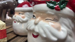 Two holiday mugs with the face of Santa on them.  The photo is up close, with one very close to the camera.  The face is winking. The second is farther back and slightly blurry.
