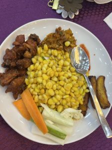 A plate of food featuring several components: crispy fried chicken pieces, a serving of corn kernels, a dollop of a yellow-orange sauce, and several pieces of fried plantains
