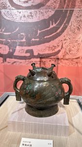 A bronze vessel with a rounded body, featuring two ornate handles and decorative designs etched into its surface. The vessel has a lid with two spouts, each topped with pointed ornaments