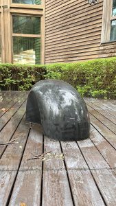 A black, curved sculpture is placed on a wooden deck, partially covered in moisture, with a background of a wooden wall and a glass window.