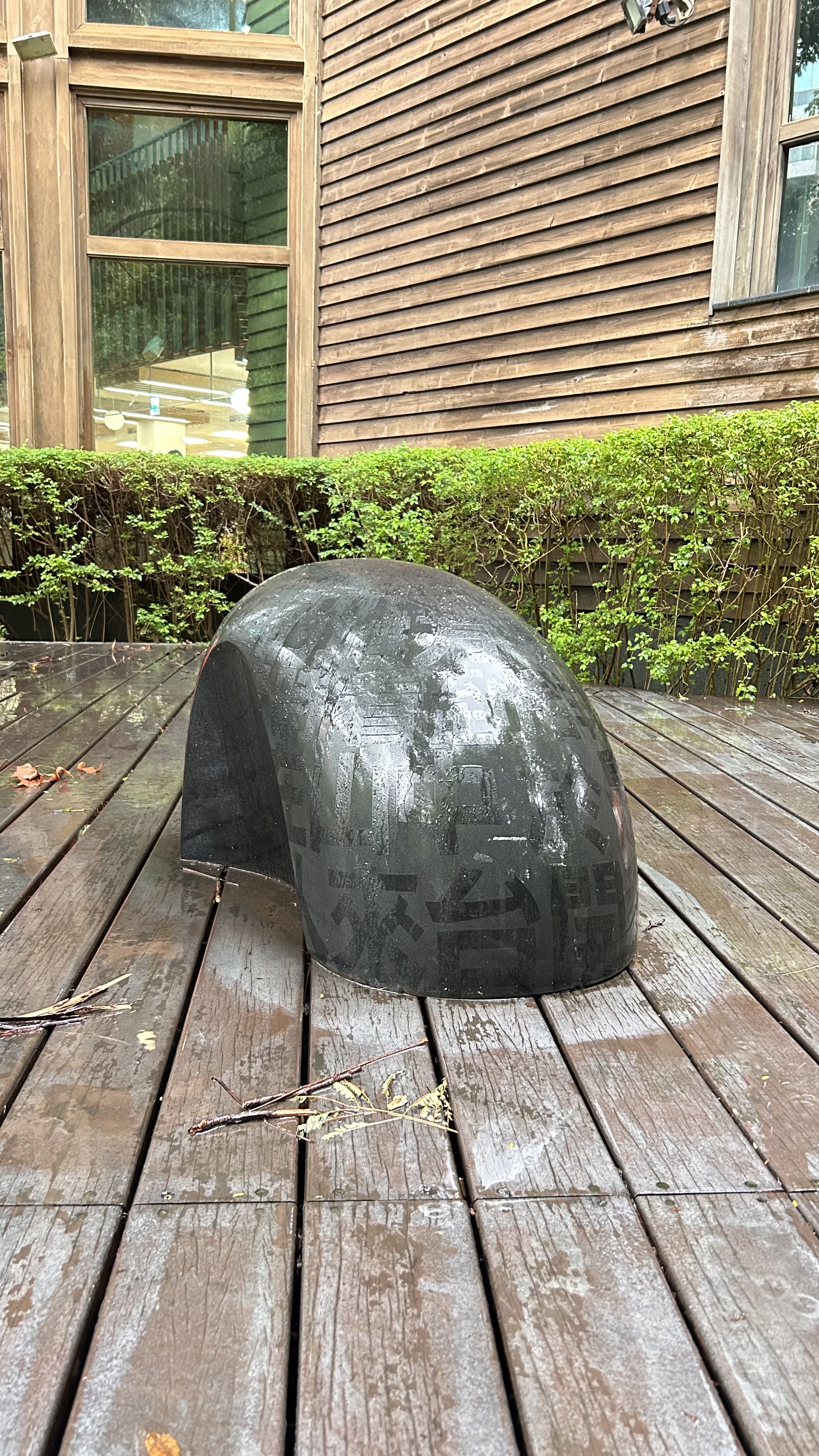 A black, curved sculpture is placed on a wooden deck, partially covered in moisture, with a background of a wooden wall and a glass window.