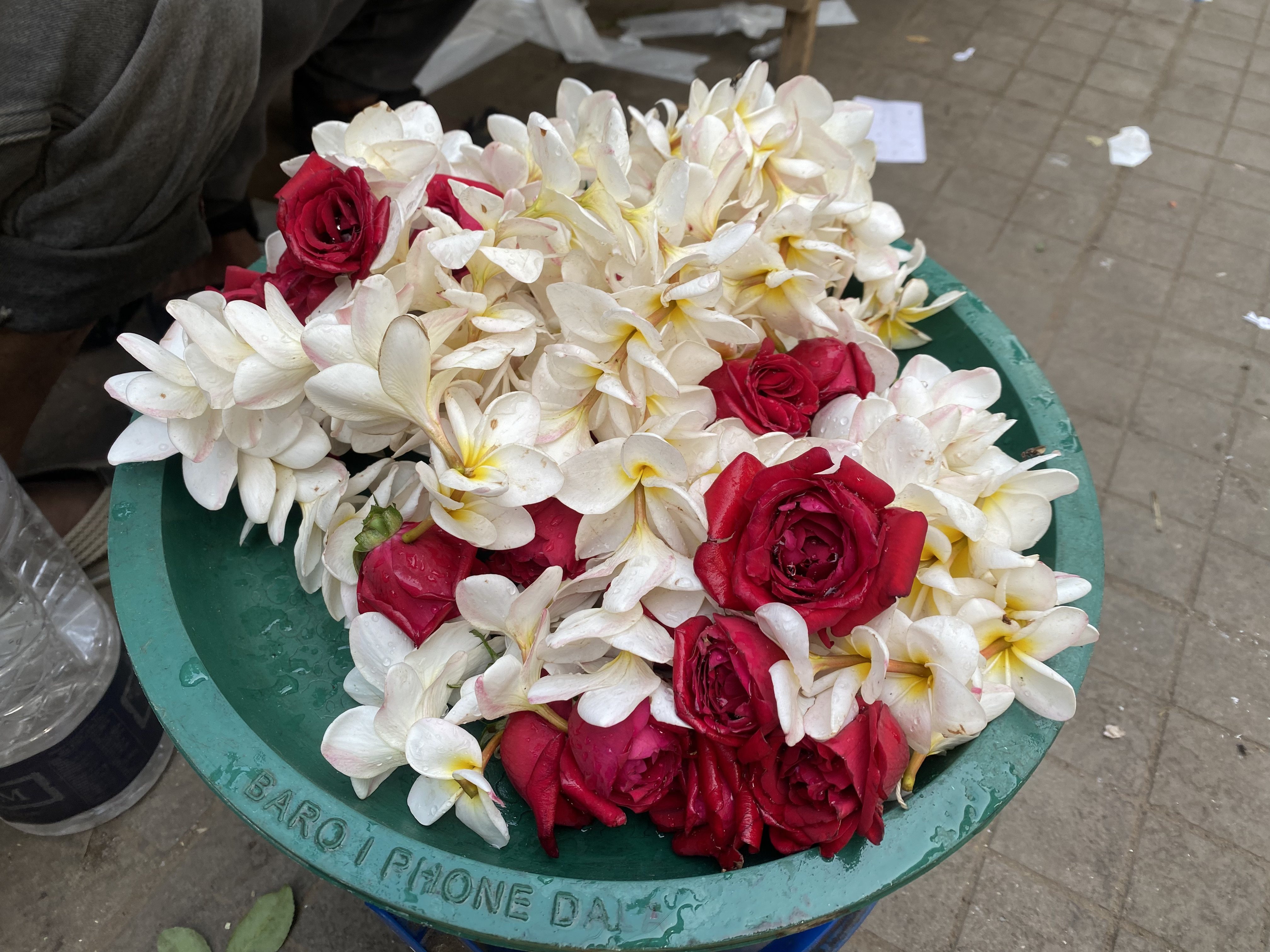 A green plastic container filled with a mix of white and red flowers, prominently featuring plumeria (frangipani) blossoms in white with yellow centers and red roses.