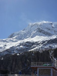 A stunning snowy mountain peak rises against a clear blue sky, with wisps of clouds drifting around the summit.