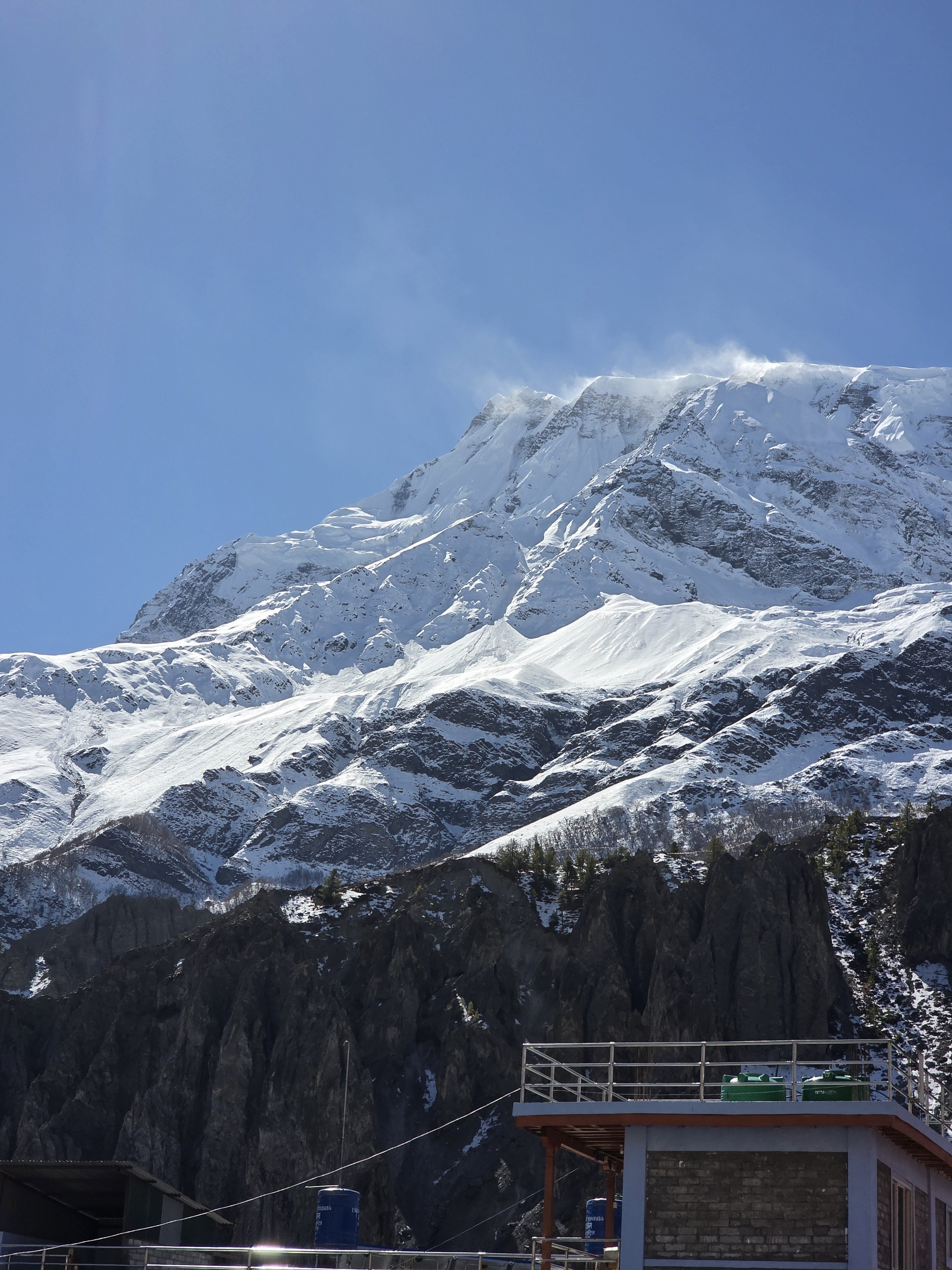 A stunning snowy mountain peak rises against a clear blue sky, with wisps of clouds drifting around the summit.
