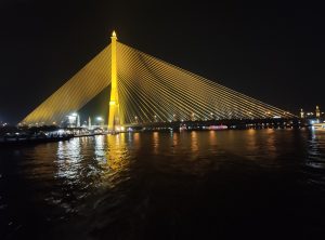 Illuminated cable stayed bridge over a river in Thailand at night.
Golden lights highlight the bridge’s tower and sweeping cables against the dark sky, while their reflections shimmer on the calm river below. Boats and distant city lights create a vibrant nighttime atmosphere.
