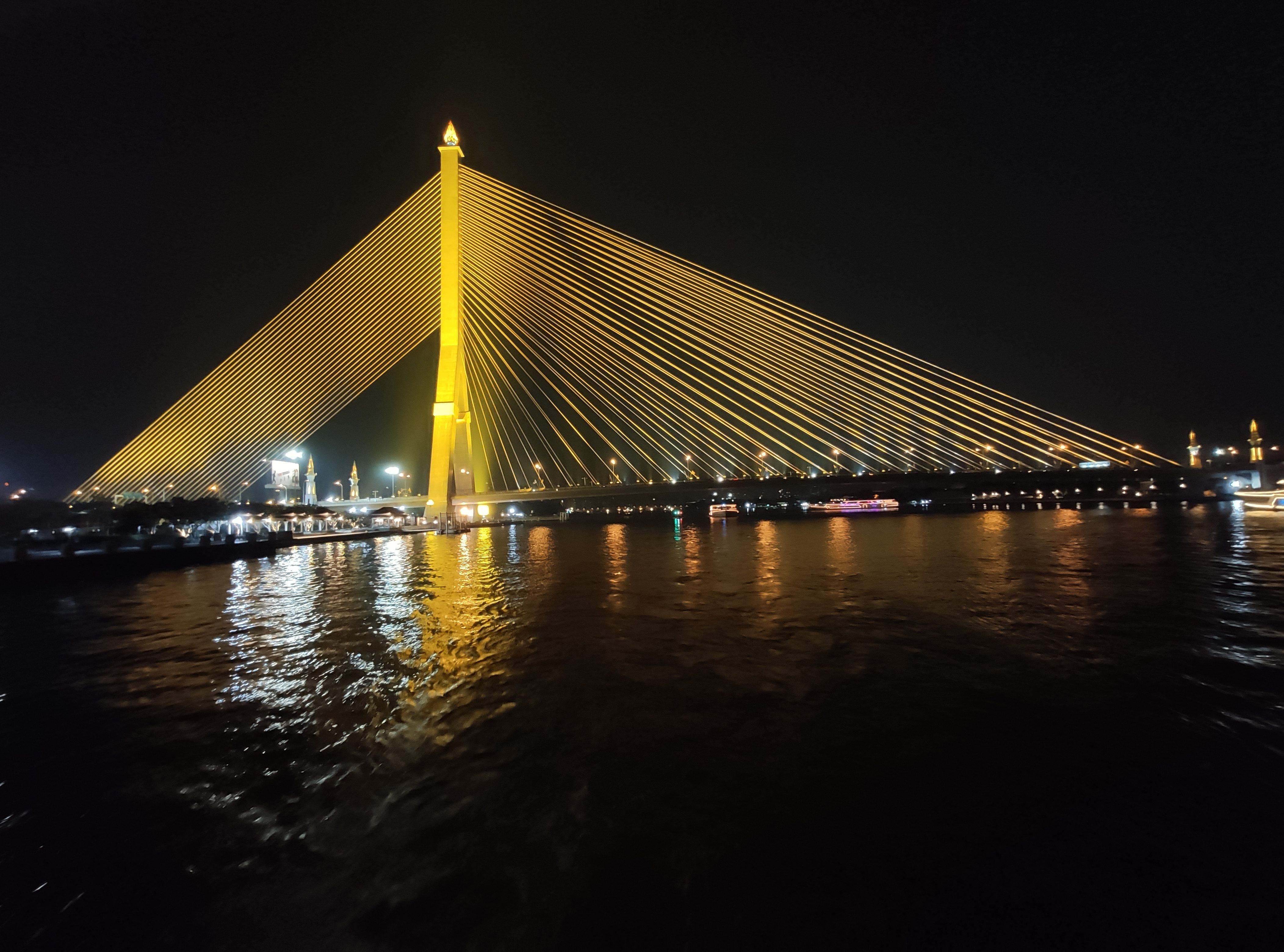 Illuminated cable stayed bridge over a river in Thailand at night.
Golden lights highlight the bridge’s tower and sweeping cables against the dark sky, while their reflections shimmer on the calm river below. Boats and distant city lights create a vibrant nighttime atmosphere.