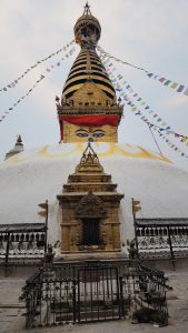 A majestic stupa adorned with golden details and prayer flags rises toward the sky, with the iconic Buddha eyes gazing outward and a calm dog resting peacefully at its base.
