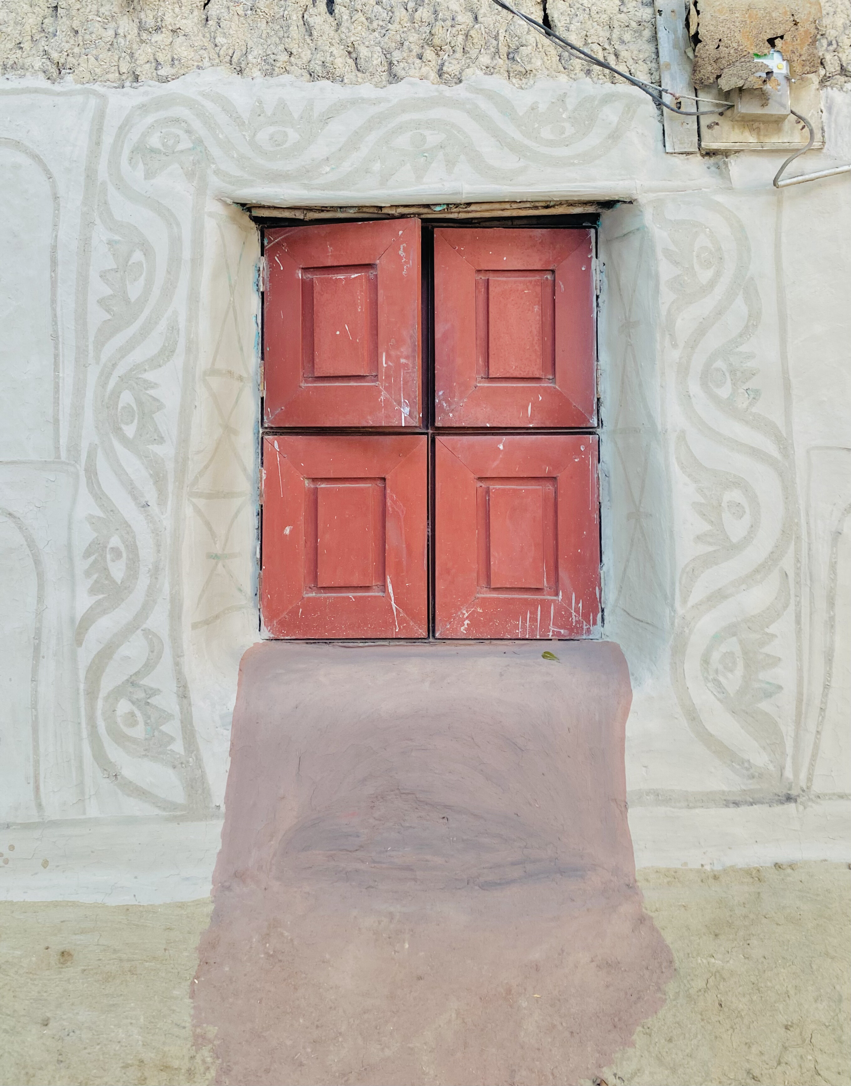 A weathered wall featuring a rectangular window adorned with four red wooden panels.