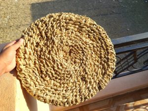 

A hand holding a round, woven straw mat (sukul) with a textured, light brown surface.