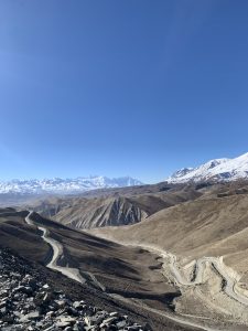 A panoramic view of a mountainous landscape featuring winding roads that carve through rocky, arid terrain. In the background, snow-capped peaks contrast with the brown hills under a clear blue sky. 