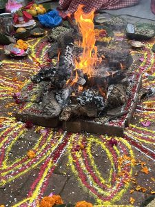 A ritual fire burns on a brick base, surrounded by elaborate circular floor patterns made of red and yellow powder and flower petals.