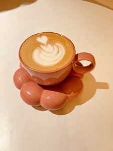 A pink cup of latte with white heart-shaped latte art, sitting on a matching pink saucer shaped like a puffy flower.