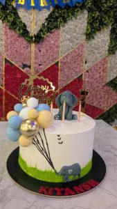 A picture of a cake featuring mini pictures of an elephant, with a 'Happy Birthday' tag placed on a white table and a multi-colored floral background.