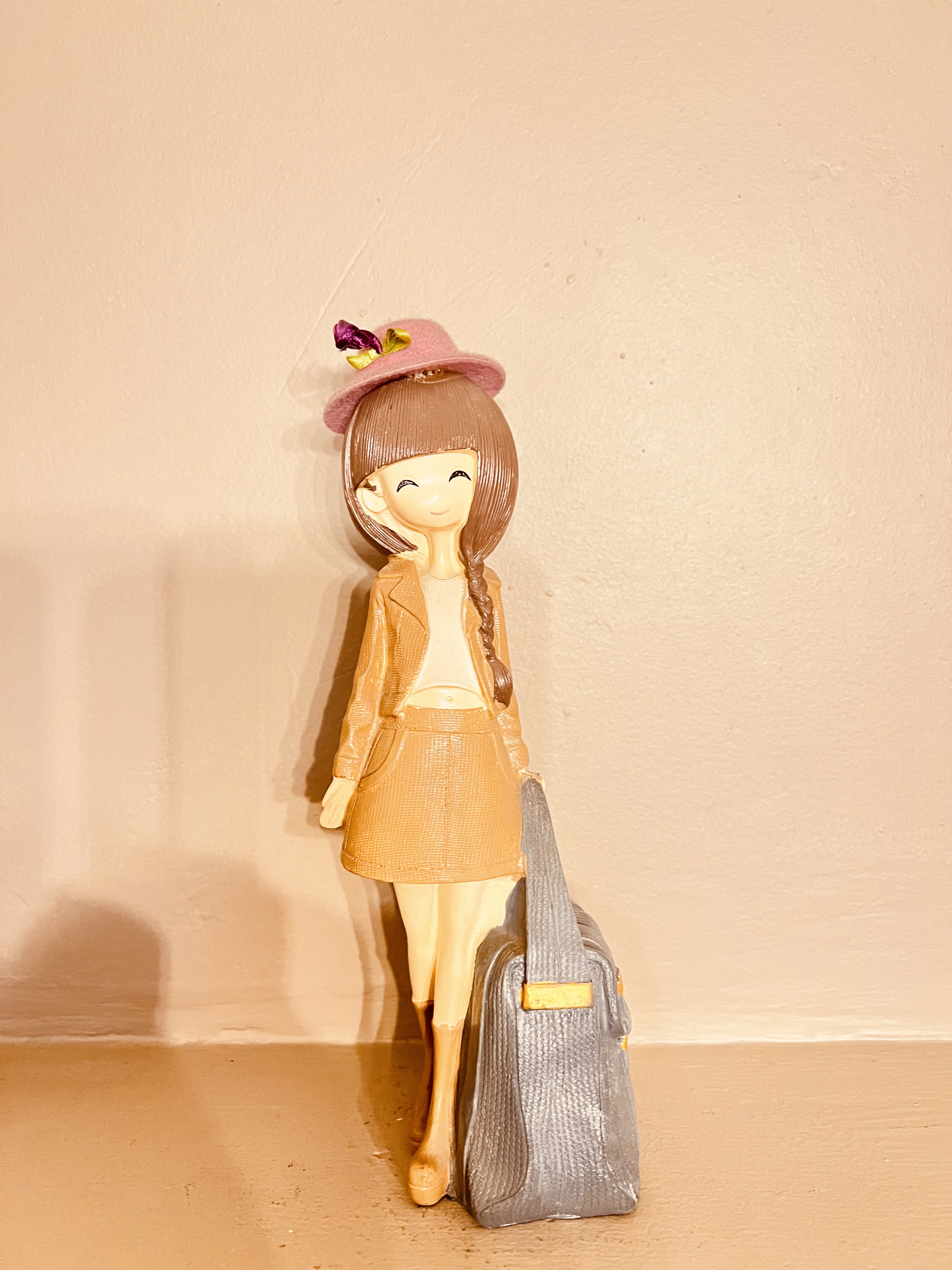 A small figurine of a smiling girl with long brown hair and a pink hat, wearing a tan outfit and standing next to a large grey suitcase.