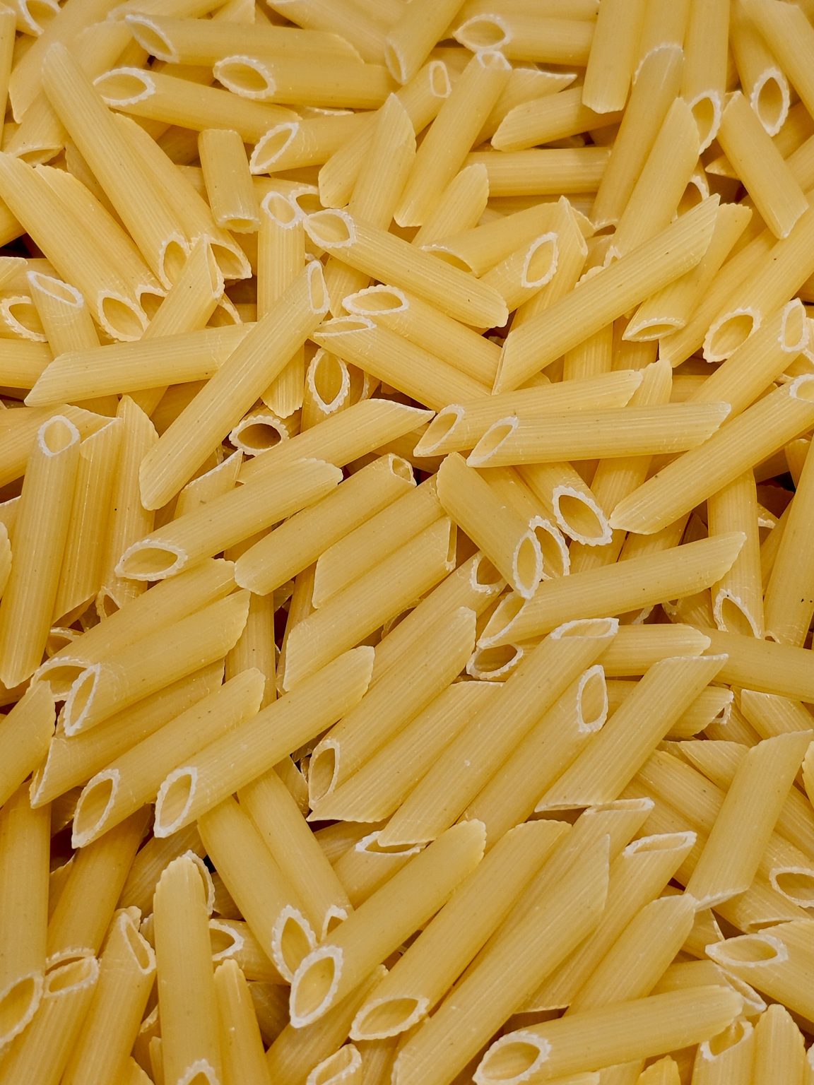 A detailed close-up of uncooked penne pasta arranged naturally, highlighting its hollow shape and smooth yellow surface. The image captures a common pantry ingredient often used in home cooking in Kozhikode, Kerala, with clean composition and sharp focus.