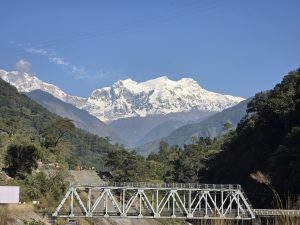 A scenic view of snow-capped mountains in the background, framed by lush green hills. In the foreground, a steel bridge spans a river or valley, with construction equipment visible on the road leading into the mountains.
