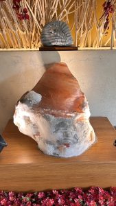 A large, irregularly shaped rock with reddish and grayish tones is displayed on a wooden surface. It is illuminated from below, casting soft light and highlighting its texture.