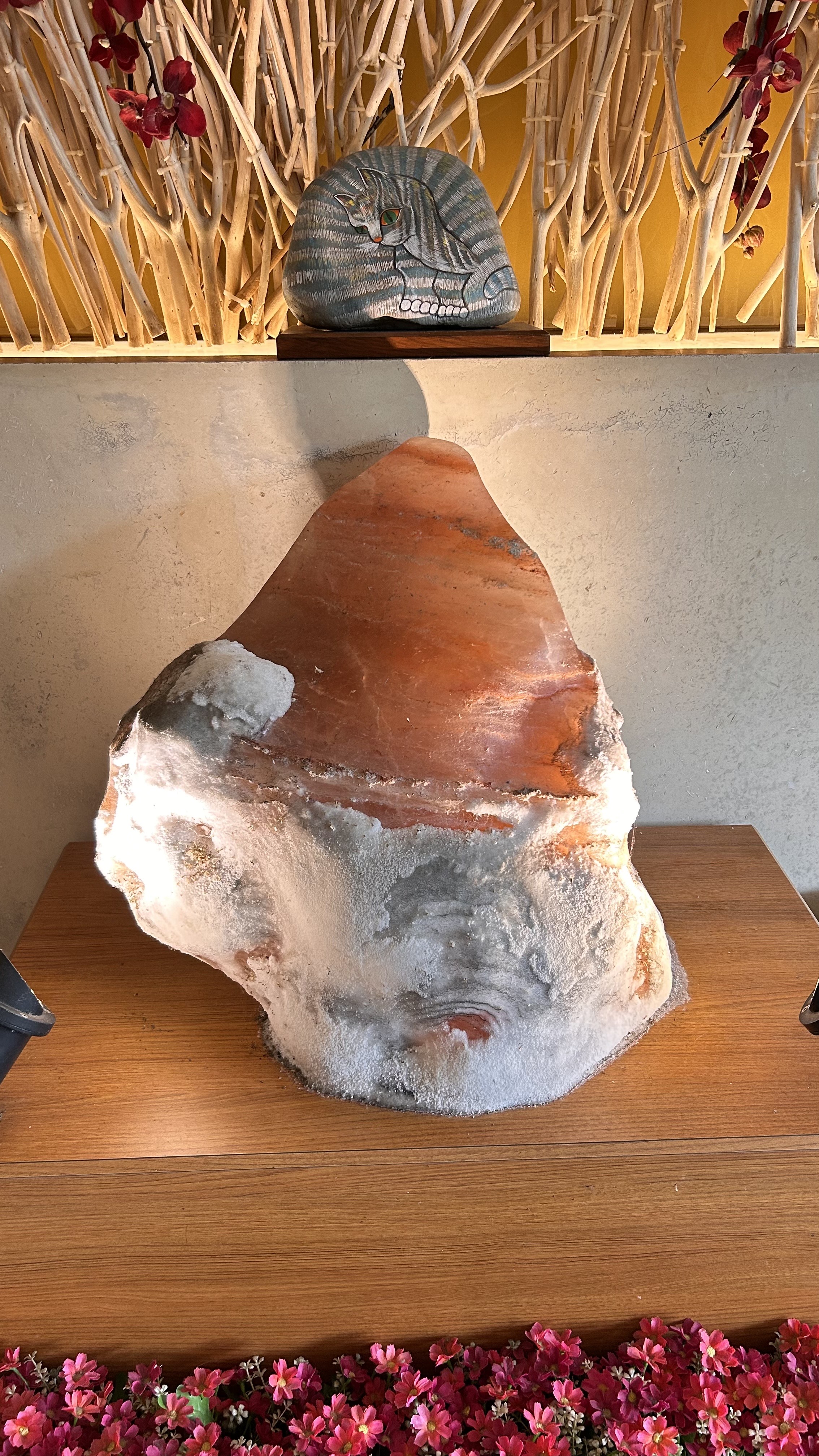A large, irregularly shaped rock with reddish and grayish tones is displayed on a wooden surface. It is illuminated from below, casting soft light and highlighting its texture.