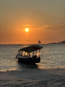 A small boat rests gently on the shoreline as the sun sets in warm golden light, casting reflections across the water with a distant volcano silhouetted on the horizon.