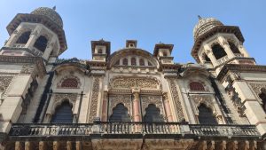 A detailed view of an ornate indo-saracenic style heritage building, featuring intricately carved stonework, arched windows, decorative balconies, and domed towers set against a clear blue sky.