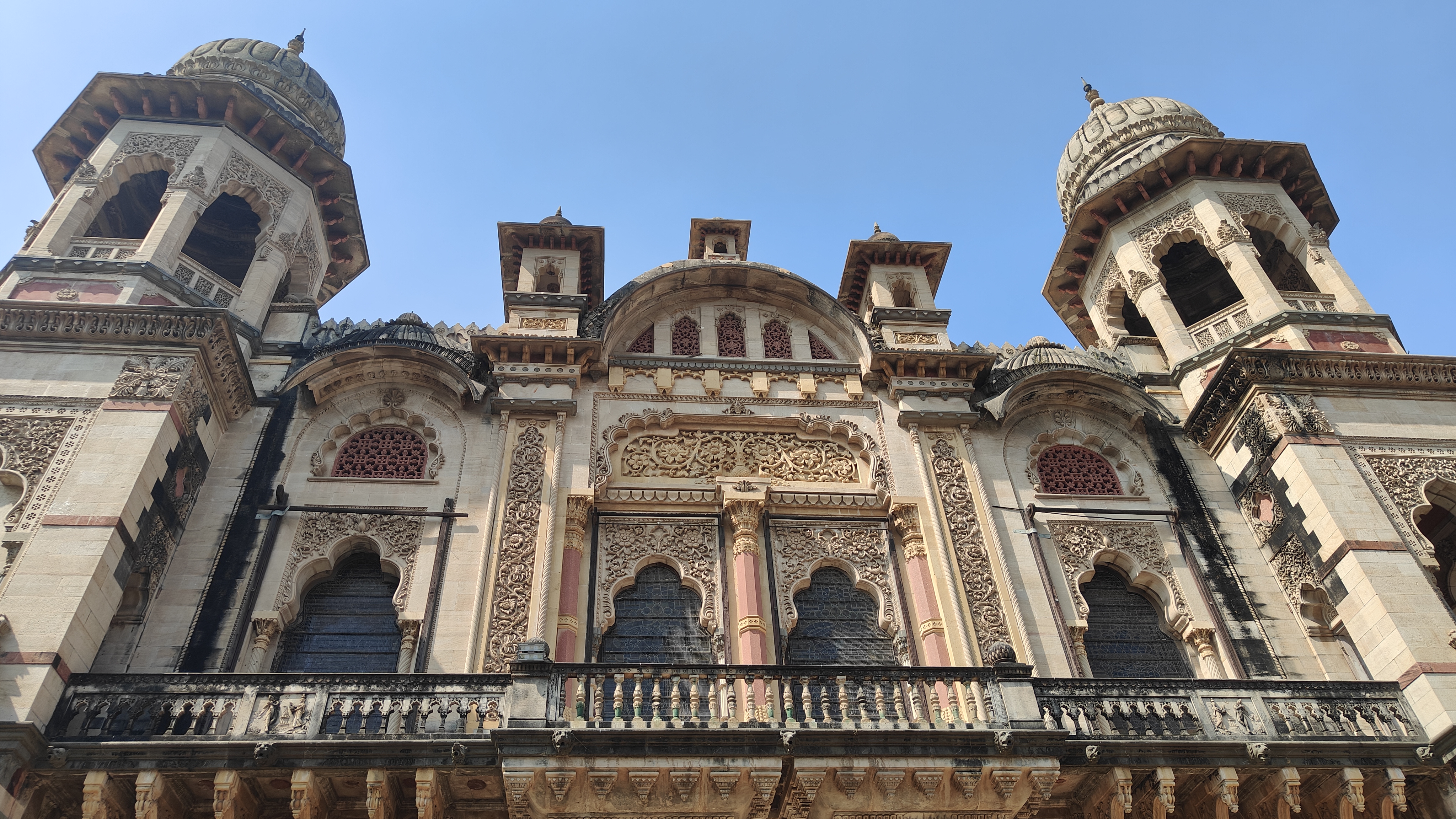 A detailed view of an ornate indo-saracenic style heritage building, featuring intricately carved stonework, arched windows, decorative balconies, and domed towers set against a clear blue sky.