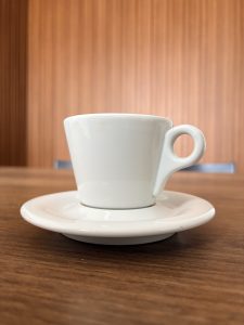A white ceramic coffee cup with a handle sits on a matching saucer, placed on a wooden table. The background is softly blurred, showing vertical wooden panels in warm tones.