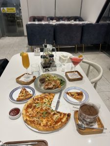 A dining table covered with various dishes, including a large pizza with toppings, a slice of pizza on the side, a plate of grilled chicken with noodles and vegetables, and a clear container of desser