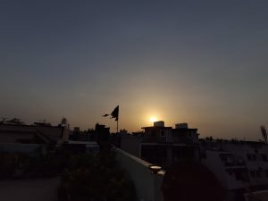 A dark silhouette of a residential skyline is framed against the warm, golden glow of a sun setting behind the buildings.