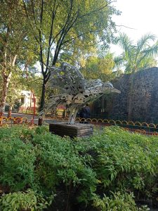 Artistic metallic sculpture of a peacock in a lush garden, crafted from intricately assembled metal pieces, perched on a stone pedestal surrounded by greenery, with trees, a stone wall, and a walking path in the sunlit background.