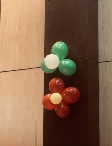 Two flower-shaped balloon arrangements, one green and white and one red and yellow, are attached to a dark brown pillar.