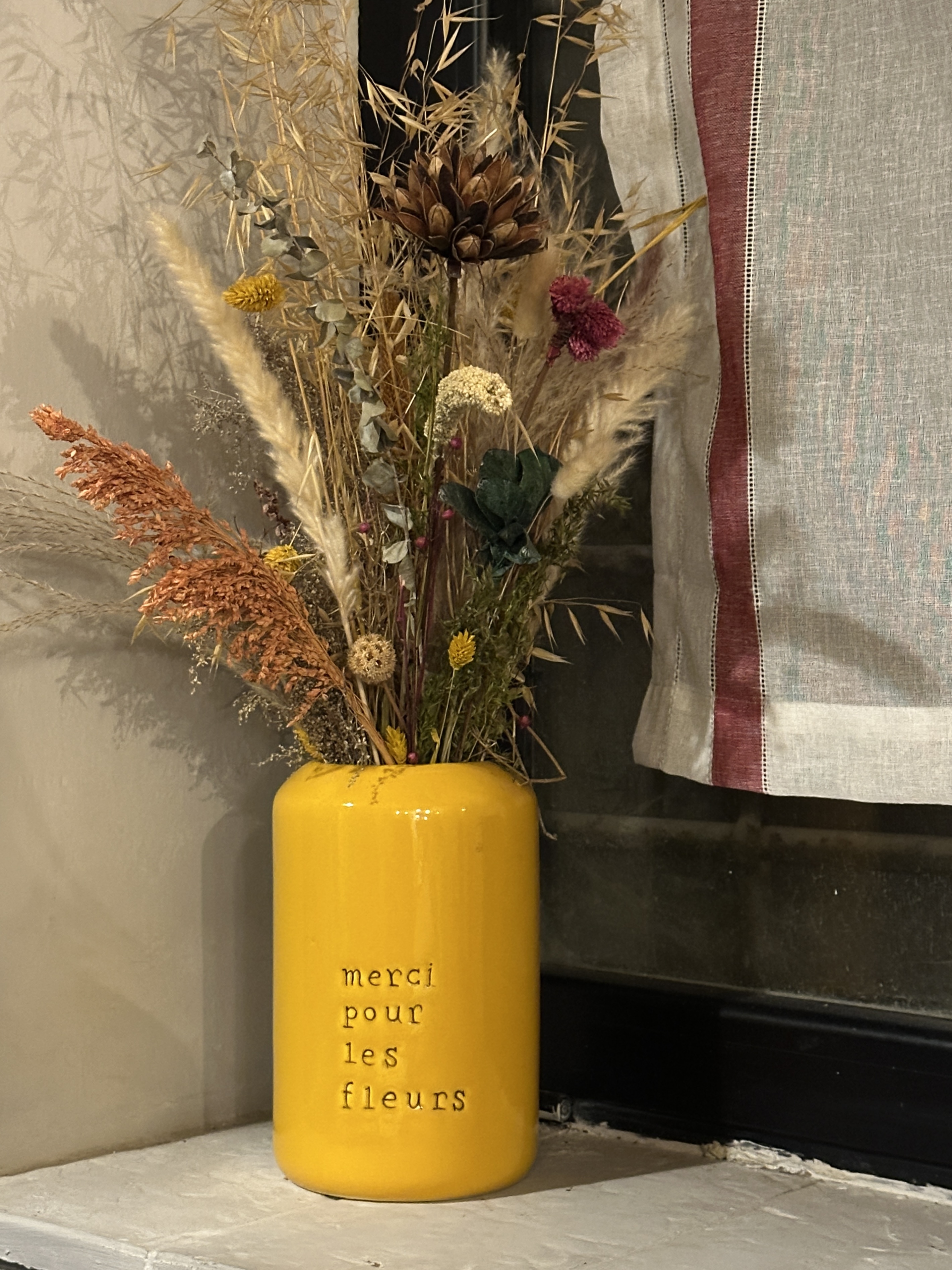 A bright yellow ceramic vase filled with dried flowers in shades of orange, yellow, green, and purple sits on a light surface near a window with a sheer curtain.