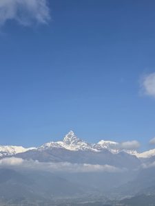 A panoramic view of a majestic snow-capped mountain range under a clear blue sky, with some wispy clouds appearing in the upper corners