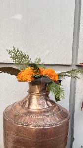 A copper pitcher adorned with vibrant orange marigold flowers, fresh green leaves, and decorative foliage sits against a light gray wall.