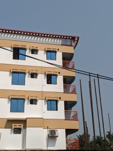 A side view of a modern multi-story building featuring white and beige painted walls, multiple blue windows, and red railings.