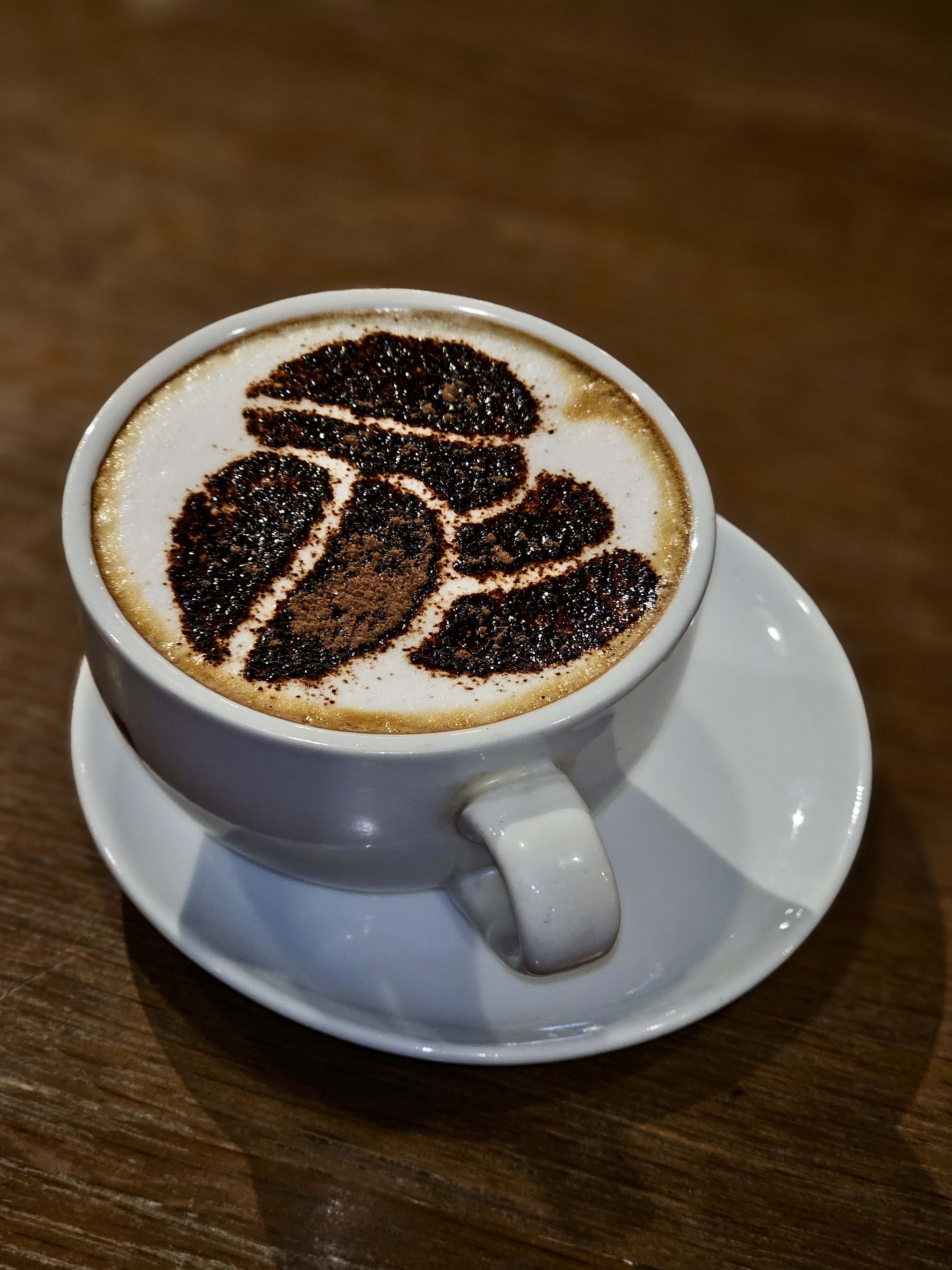 A freshly prepared cup of coffee served in a white ceramic cup, topped with cocoa powder art on smooth milk foam. Captured indoors in Kozhikode, Kerala, this image reflects a calm café moment with warm lighting and a cozy feel.