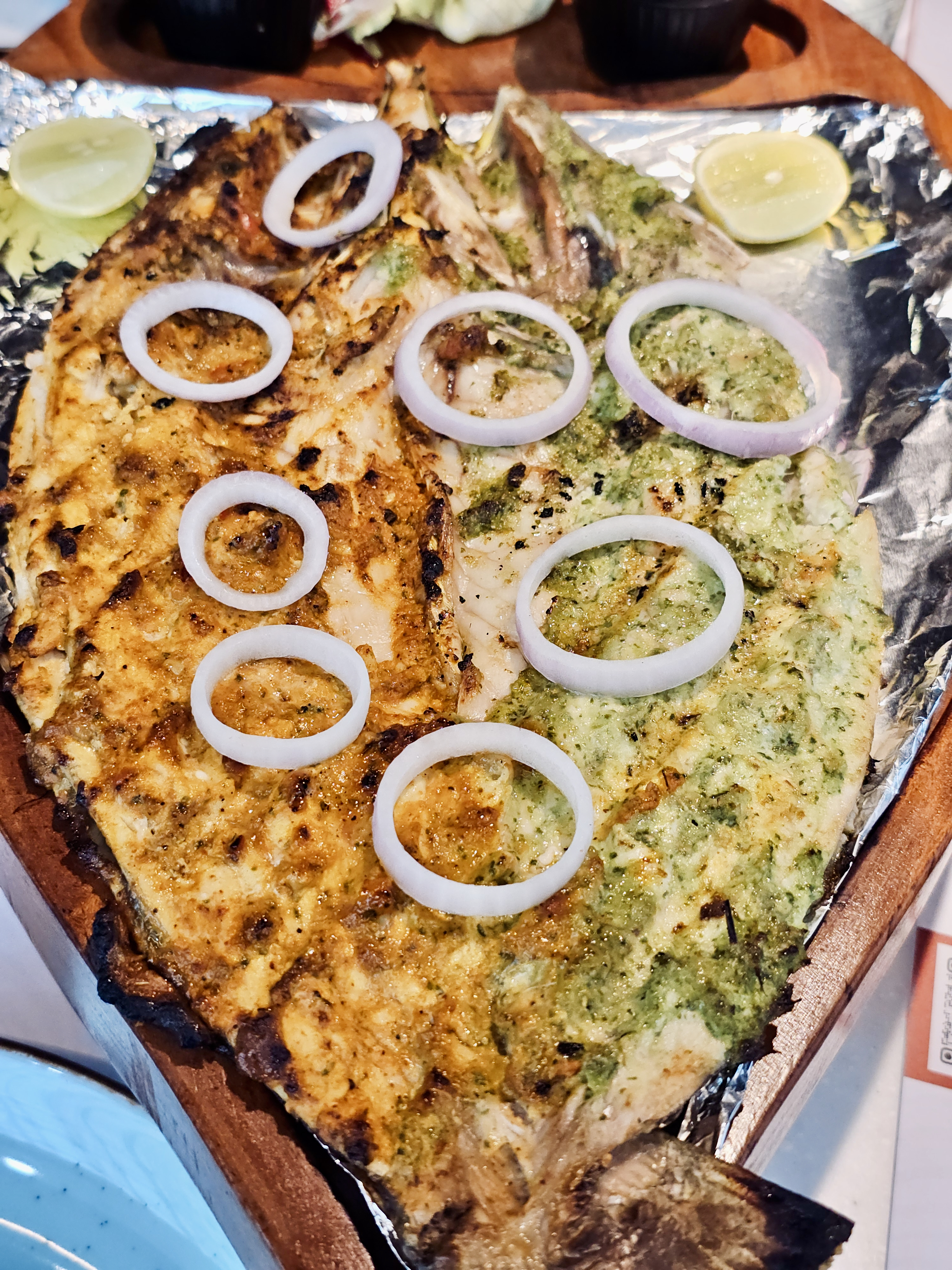 A large grilled fish served with green and red marinades, topped with onion rings and lemon slices. The overhead view shows vibrant colors and appetizing details, photographed at a restaurant in Kozhikode, Kerala. 