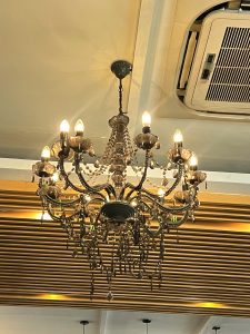 A large, ornate vintage chandelier with numerous bronze arms, frosted shades, and hanging crystals is suspended from a ceiling above wooden slat paneling.