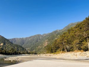 A scenic view of a riverbank surrounded by lush green mountains under a clear blue sky