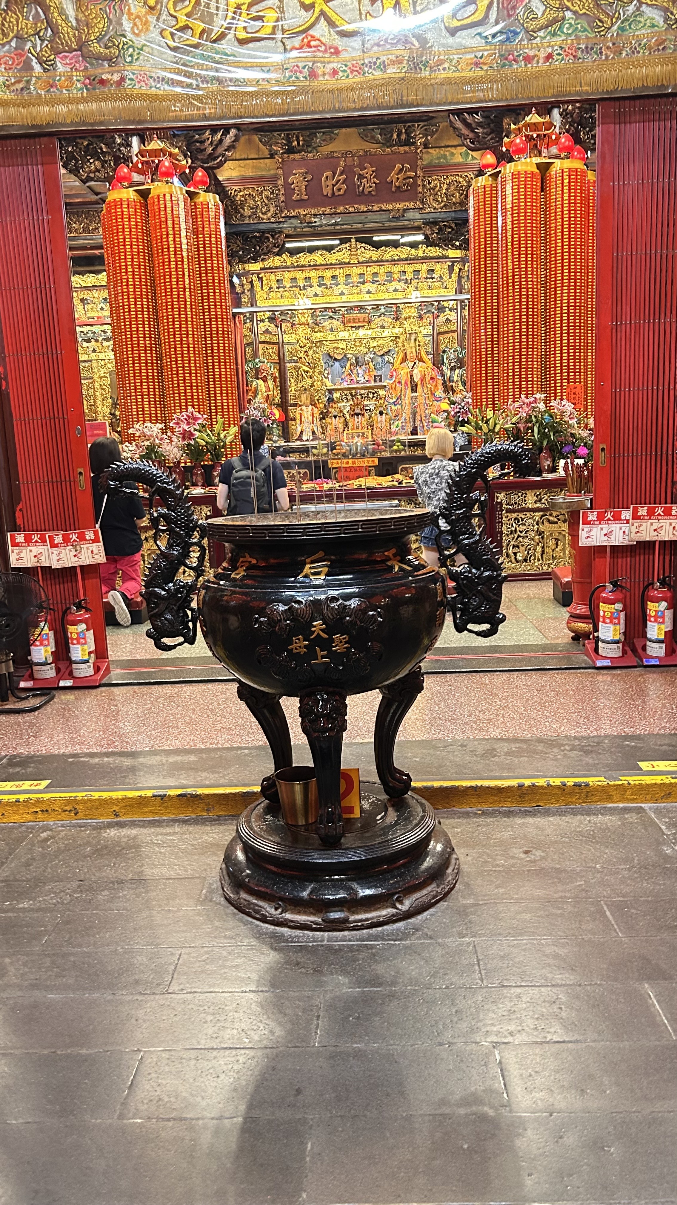 A black incense burner with dragon motifs stands prominently in a vibrant temple interior. 