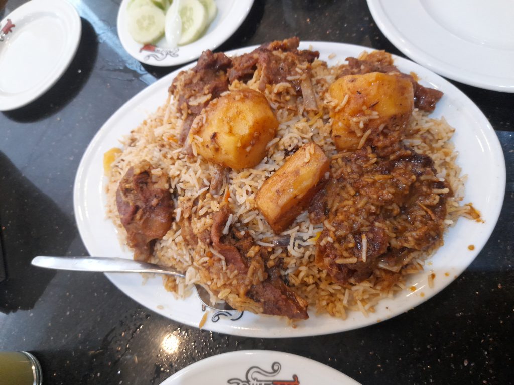 A plate of biryani with meat, potatoes, and cucumber on a dark table in Harbaid, Gazipur.
