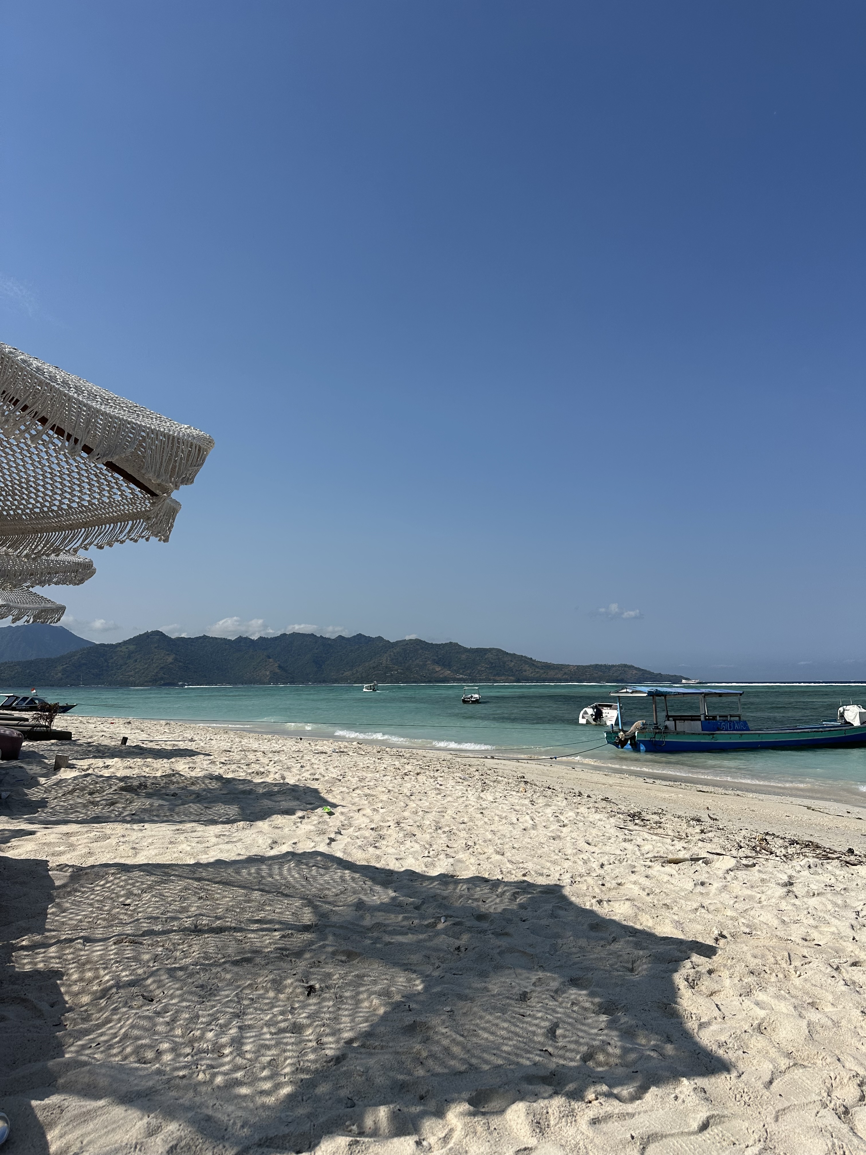 A calm tropical beach stretches out with soft white sand, turquoise water, and distant green mountains under a perfectly clear blue sky.