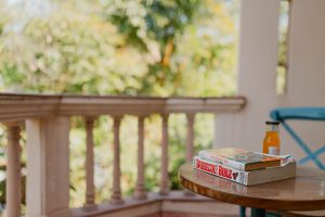 This image portrays a quiet, leisurely moment on a shaded balcony or veranda. In the foreground, a small round wooden table holds a short stack of books and a bottle of orange-colored drink, suggesting a pause for reading and refreshment. The table’s polished surface reflects soft daylight, adding warmth to the scene.